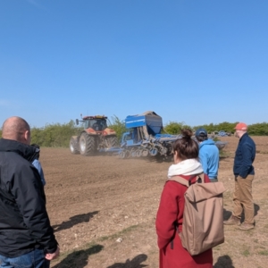 Attendees standing in a field, watching a tractor, and hearing a talk about FAI Farm.