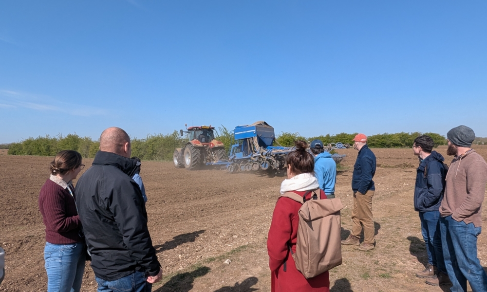 Attendees standing in a field, watching a tractor, and hearing a talk about FAI Farm.